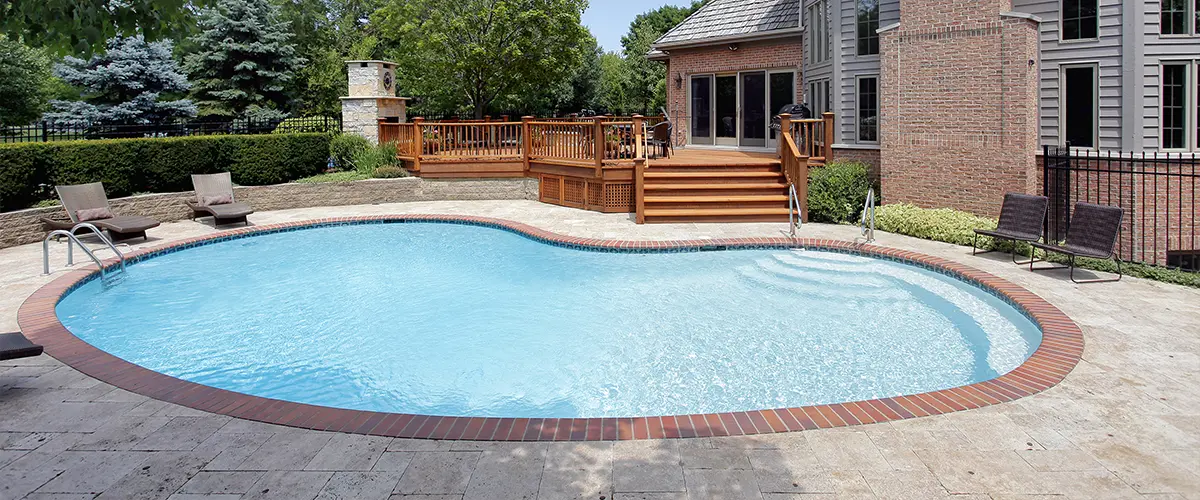 Beautiful backyard in Little Elm, with an inground swimming pool, wooden deck, lounge chairs, outdoor fireplace, and a large brick house surrounded by lush greenery.