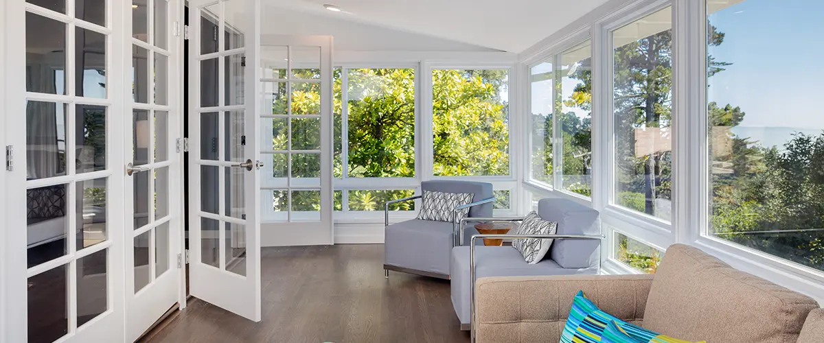 Bright sunroom in Frisco with large windows, modern furniture, and French doors opening to a scenic view of lush greenery and blue sky