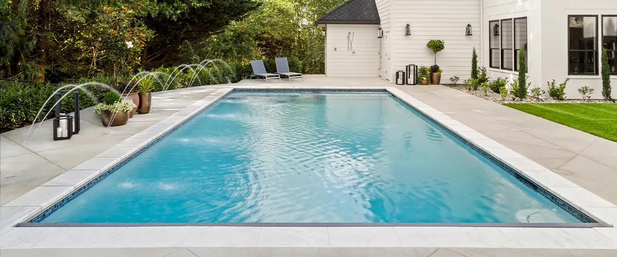 Modern backyard in Little Elm, with an inground swimming pool, water features, lounge chairs, potted plants, and a white house surrounded by lush trees.