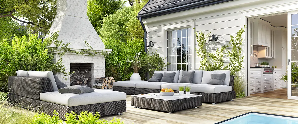 Modern outdoor patio in Frisco, with comfortable outdoor furniture, a white brick fireplace, lush greenery, and access to a stylish indoor kitchen through sliding glass doors.