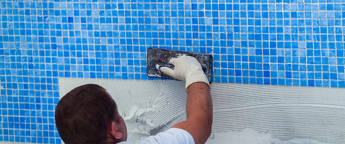 Worker doing a pool repair job with blue mosaic tiles using a trowel and adhesive, wearing gloves for protection.