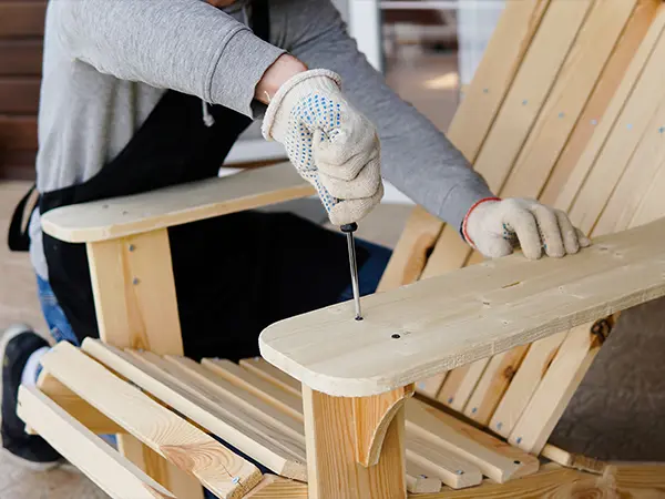 Person wearing gloves assembling a wooden outdoor chair with a screwdriver, focusing on the armrest and slatted seat.