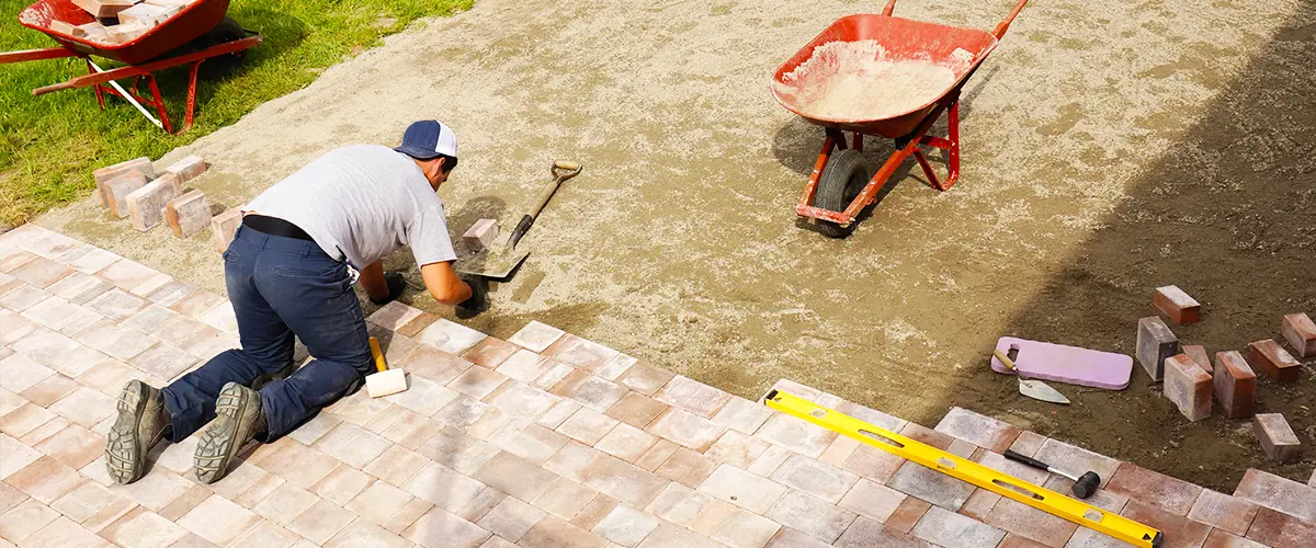 Construction worker installing pavers for a patio with tools, red wheelbarrow, bricks, and a partially completed stone layout.