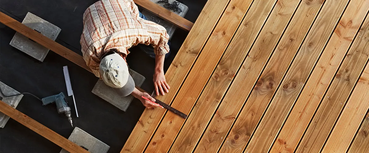 Worker installing wooden deck boards with measuring tools and drill - construction of outdoor patio decking.