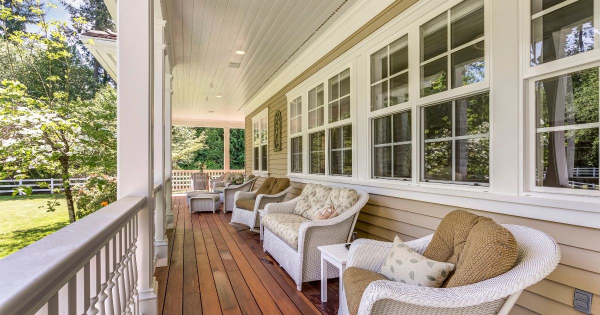 A classic covered porch with warm wood flooring, white wicker seating, and plush cushions, overlooking a green yard with trees and a white fence