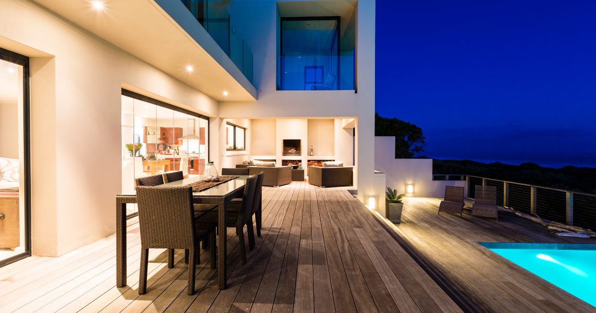 A modern outdoor patio at night with wooden decking, stylish seating, ambient lighting, and a poolside view