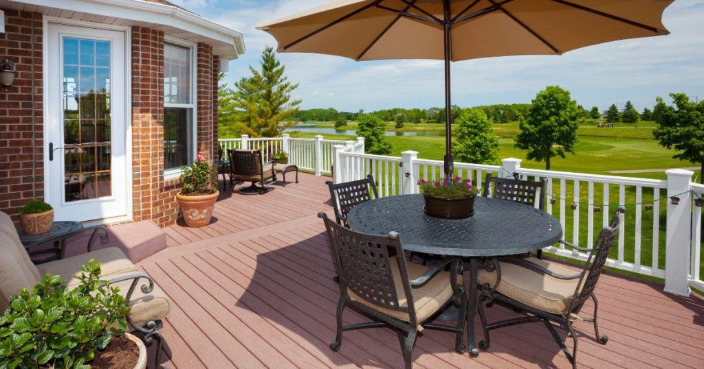 A composite deck attached to a brick house, featuring black wrought iron patio furniture with beige cushions, a large umbrella, and a white railing overlooking a lush green golf course