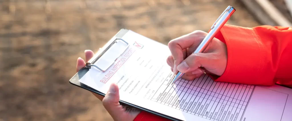 A building inspector in a red jacket filling out a building inspection report on a clipboard at an outdoor site