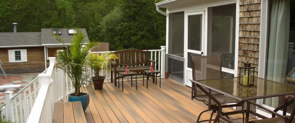 A cozy deck with potted plants, a wooden bench, and a glass-top table, featuring a white railing and natural wood siding