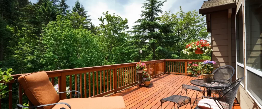 A cozy wooden deck with metal patio furniture, a cushioned lounge chair, and vibrant flower planters, surrounded by lush green trees