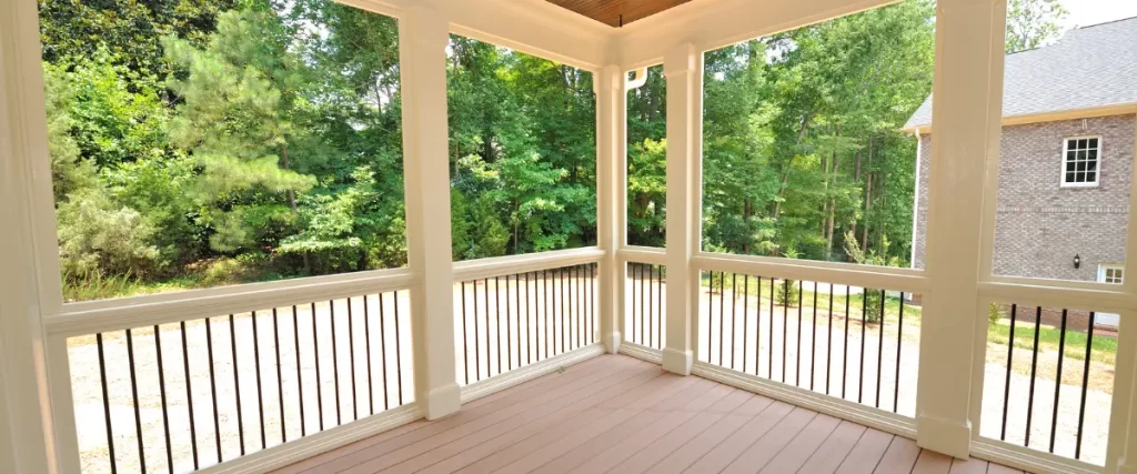 An enclosed porch with white trim, a brown ceiling, and a composite deck floor, offering a view of surrounding trees