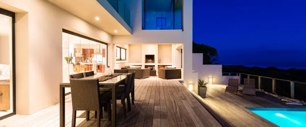 A luxurious outdoor patio with wooden decking, a modern fireplace, outdoor dining, and a pool overlooking a scenic view at dusk