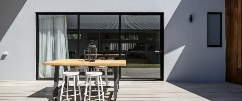 A minimalist deck with a modern wooden bar-height table and white stools, attached to a contemporary home with sliding glass doors