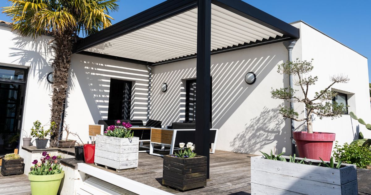 A stylish outdoor patio with a sleek black pergola casting striped shadows on a white modern home, complemented by vibrant potted plants and contemporary seating."