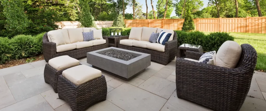  Outdoor patio with a modern fire pit and wicker furniture set, surrounded by lush greenery.