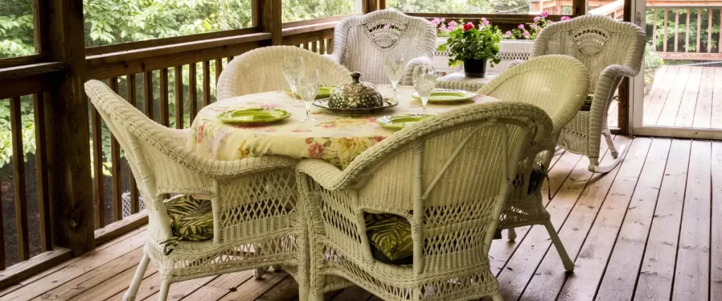 A screened-in porch with a rustic wooden floor, featuring a white wicker dining set with floral cushions and a decorative table setting