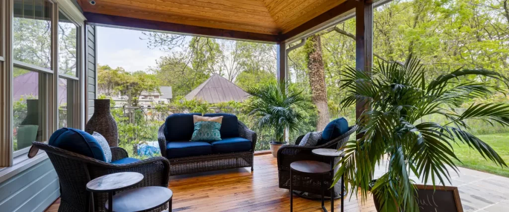 A screened-in porch with wooden floors, dark wicker seating with blue cushions, and tropical plants, creating a relaxing outdoor space