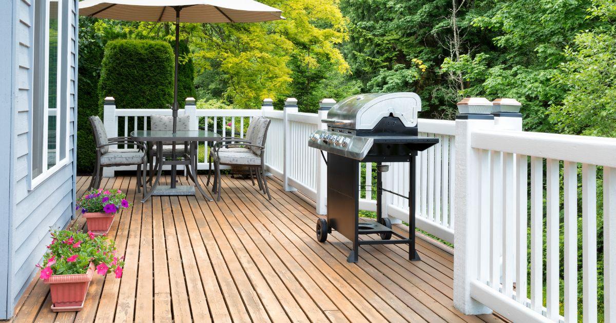 A wooden deck with a black grill, patio dining set, and a white railing, attached to a house with greenery in the background