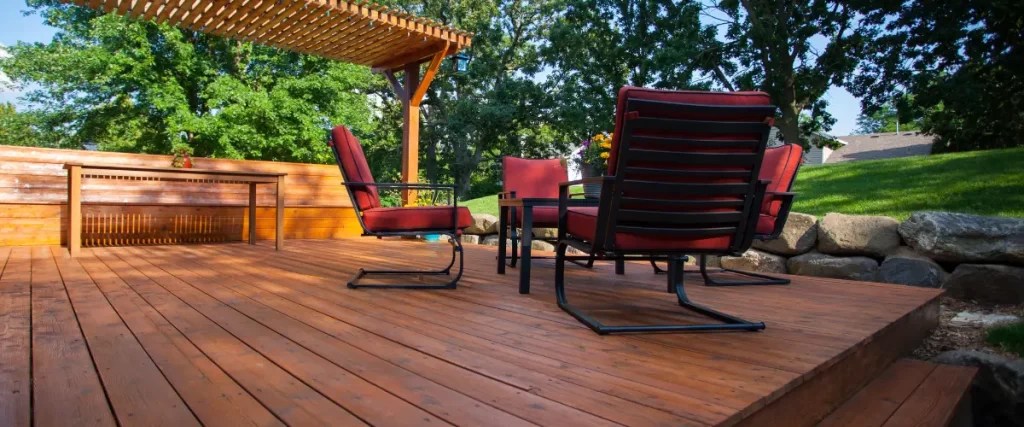 A wooden deck with a black grill, patio dining set, and a white railing, attached to a house with greenery in the background